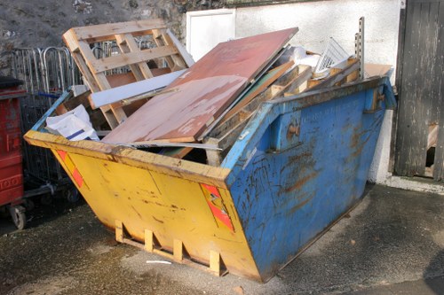 A large blue and yellow industrial rubbish skip filled with a mix of discarded building materials and waste, including wooden pallets, metal sheets, and various pieces of broken timber, situated on an outdoor paved area adjacent to a concrete wall and black wooden fencing. The skip's surface shows signs of weathering with rust and dirt, and it is positioned in close proximity to other waste management equipment. The background includes part of a dark-colored gate and a section of white wall. The scene is well-lit, indicating daytime, and the overall environment suggests a storage or disposal area typical for waste removal services in the Acton area, with the company Business Waste Removal Acton handling the collection and management of rubbish.