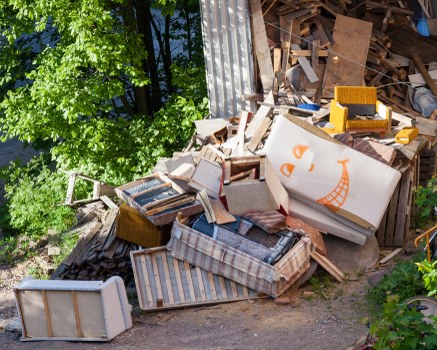 A large pile of mixed waste materials is shown outdoors, positioned against a corrugated metal fence. The collection includes wooden furniture pieces such as a striped upholstered armchair, a small wooden table, and a wooden bench, all with visible wear and damage. There are also cardboard boxes, some decorated with simple orange and white graphic designs, and various fragments of wooden planks and panels. The objects are arranged haphazardly on a paved area adjacent to lush green foliage and trees, indicating a garden or backyard setting. Bright natural sunlight illuminates the scene, casting shadows across the debris. This image relates to rubbish removal services in Acton, as provided by Business Waste Removal Acton, who handle the collection and disposal of such household and garden waste materials, typical of a local rubbish clearance operation.