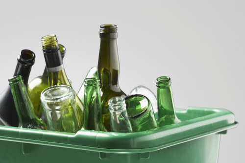 A green plastic bin filled with various clear and dark glass bottles, some with metal caps and others open, positioned on a flat surface against a plain light background. The bottles are of different heights and shapes, with some leaning to the side and others standing upright. The bin's edges are smooth, and the bottles appear clean, ready for disposal or recycling. This image reflects typical waste collection for glass bottles, relevant to rubbish removal services in Acton, London, such as those offered by Business Waste Removal Acton. The setting is simple and uncluttered, focusing solely on the bottles and the bin, illustrating the need for proper waste management of glass containers, often collected during local rubbish removal operations.
