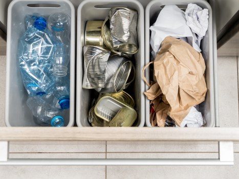 A three-compartment recycling bin placed on a tiled floor, each section filled with different types of waste. The left compartment contains several clear plastic bottles with blue caps, some crushed and others intact, indicating they are used beverage containers. The middle compartment holds a stack of metallic cans, primarily aluminium, with some crushed and others slightly dented, used for soft drinks or food products. The right compartment contains crumpled paper and a brown paper bag, both loosely folded and showing creases, suitable for general paper waste. The bins are made of light-colored plastic and are situated outdoors or in a commercial waste collection area, with the tiled floor extending underneath. The scene is well-lit, emphasizing the different textures and colors of the waste materials, which reflects proper waste sorting practices often seen in waste removal services like Business Waste Removal Acton, serving the local postcode area.