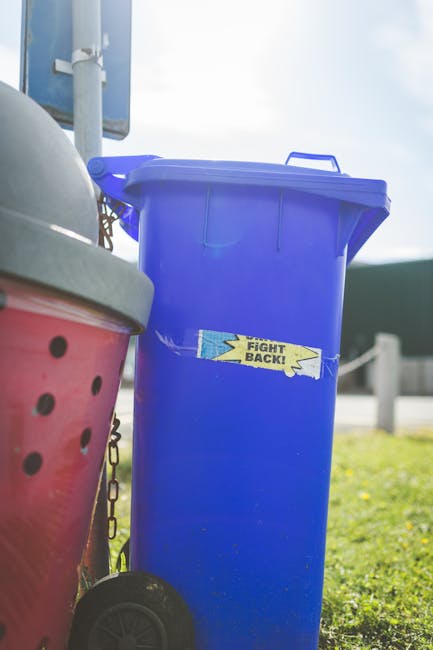 A person placing a crumpled brown paper bag into a grey rubbish bin with a white lid, which is positioned on a wooden surface. The individual's hand is visible, holding the paper bag by its top edges,