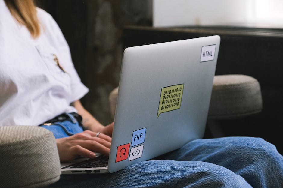 A close-up photograph of a laptop computer with a visible code editor on the screen, displaying lines of programming code in various colors, situated on a wooden desk. To the right of the laptop, ther